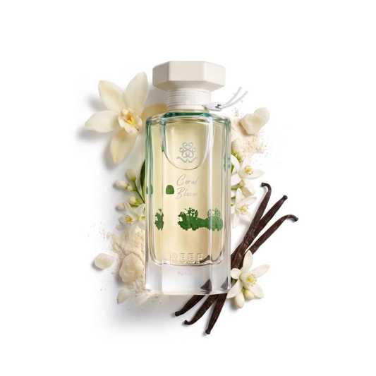 A clear glass perfume bottle with pale yellow liquid and green botanical illustrations, topped with a light beige cap. The bottle is styled with white flowers, vanilla beans, and powder on a white background.