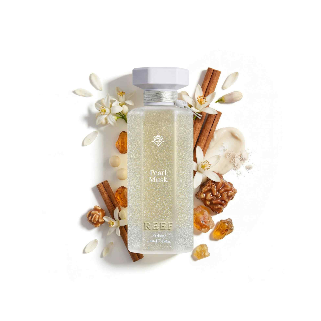 A frosted glass bottle labeled 'Pearl Musk' by REEF, filled with a pale shimmering liquid and topped with a white cap, styled on a white background with cinnamon sticks, white flowers, nuts, and amber pieces around it.